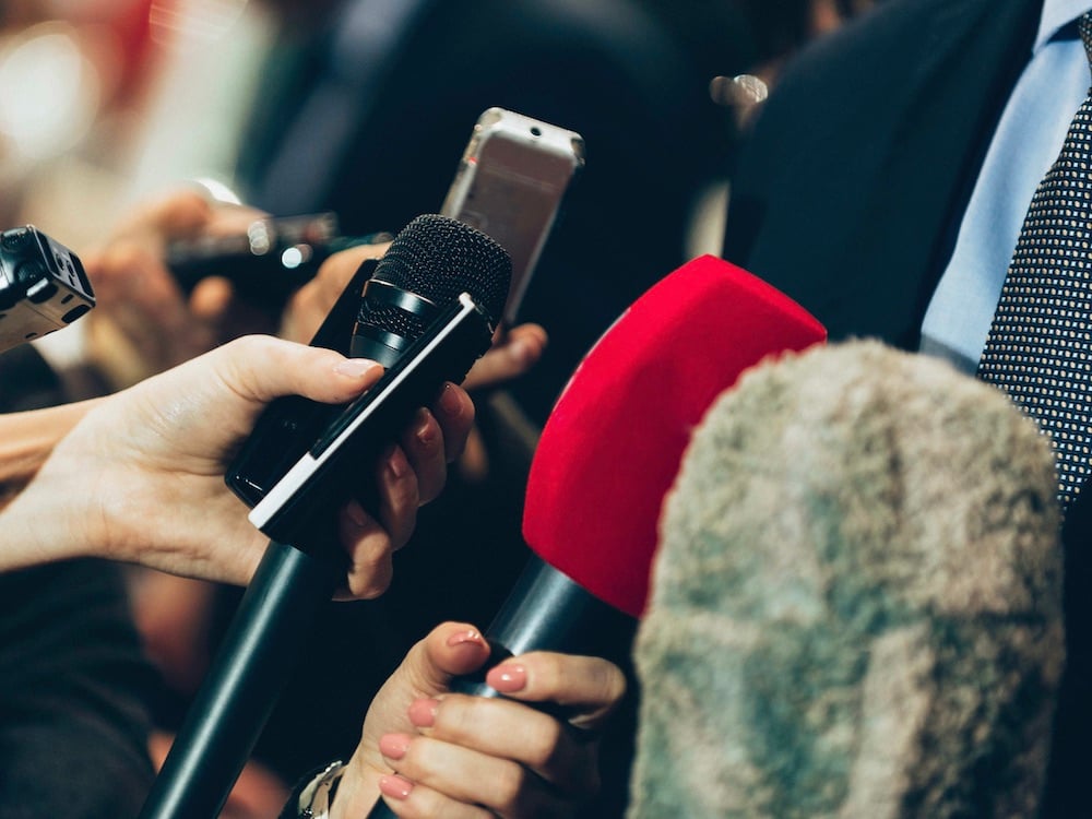 A crowd of hands holding microphones and voice recorders tilts their devices towards the right side of the screen, where a person in a navy jacket, blue shirt and dark dotted tie is standing. The hands belong to journalists in a press scrum. We can’t see their faces. 