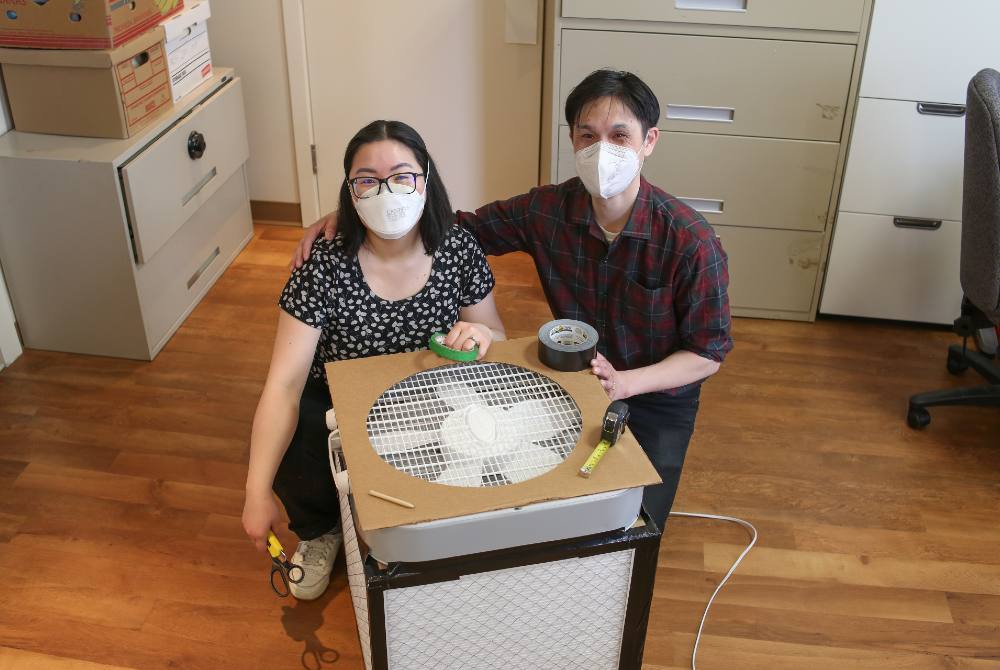 Kimberly Sayson and Jeffery Chong kneel behind a finished Corsi-Rosenthal box in an office setting with hardwood floors.