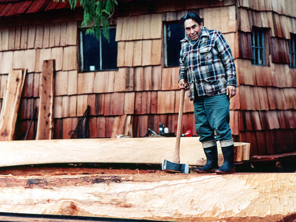 A man stands in front of a cedar cabin wearing a blue plaid jacket and holding a large axe. He’s standing on a log which is being carved into a canoe using a single piece of wood.