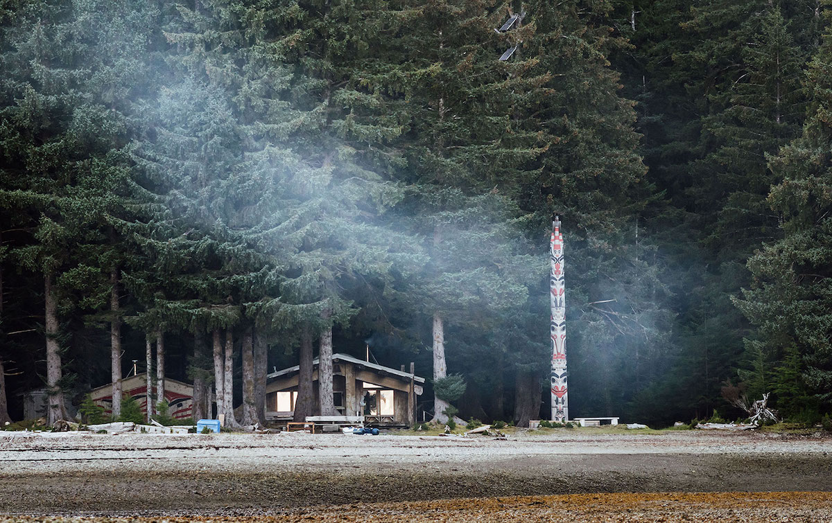 Two Haida longhouses can be seen built just behind where the beach changes into the forest. A totem pole stands, facing the seaweed-scattered beach and an unseen fire spits out a blue trail of smoke.