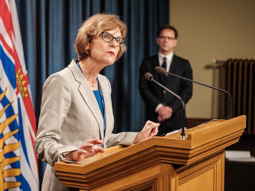 A middle-aged light-skinned woman stands at a podium, with brownish hair cut above her shoulders. She wears dark-rimmed glasses, an oatmeal jacket, a blue shirt and pearls.