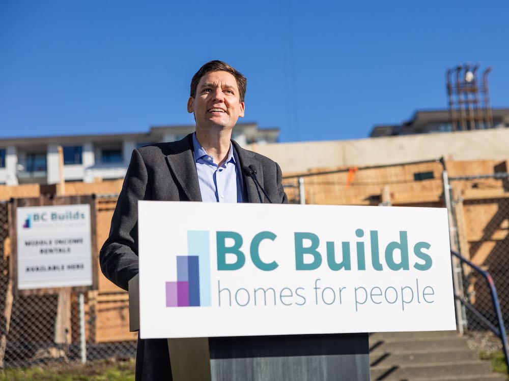 A middle-aged white man in a dark suit with no tie smiles at the camera standing at a podium with a sign that says 'BC Builds homes for people.' In the background, under blue skies, are homes under construction.