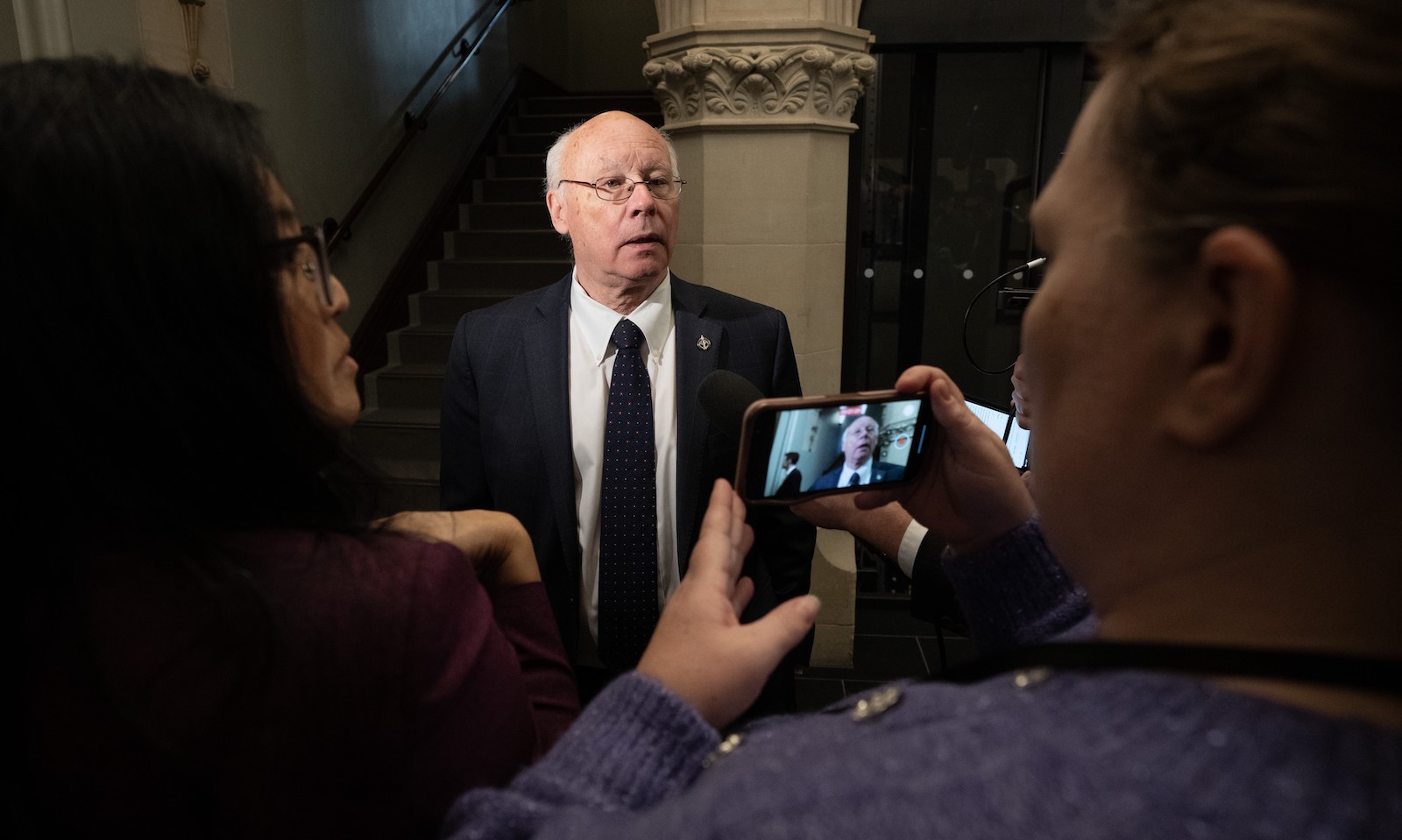 Ken Hardie has light skin, white hair and glasses. He wears a dark suit and is standing in front of a Corinthian column while a scrum of reporters interview him. In the foreground, a reporter in a blue sweater is making a video recording of Hardie on a smartphone.