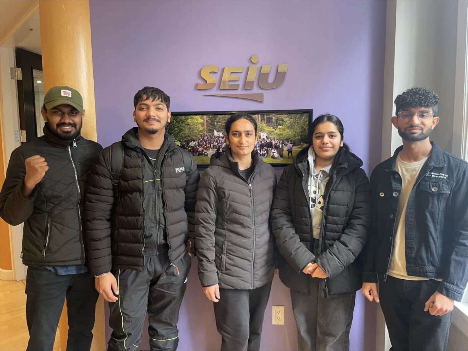 Five people in dark winter jackets stand in front of a purple wall. The wall is mounted with a metallic sign that says “SEIU.”