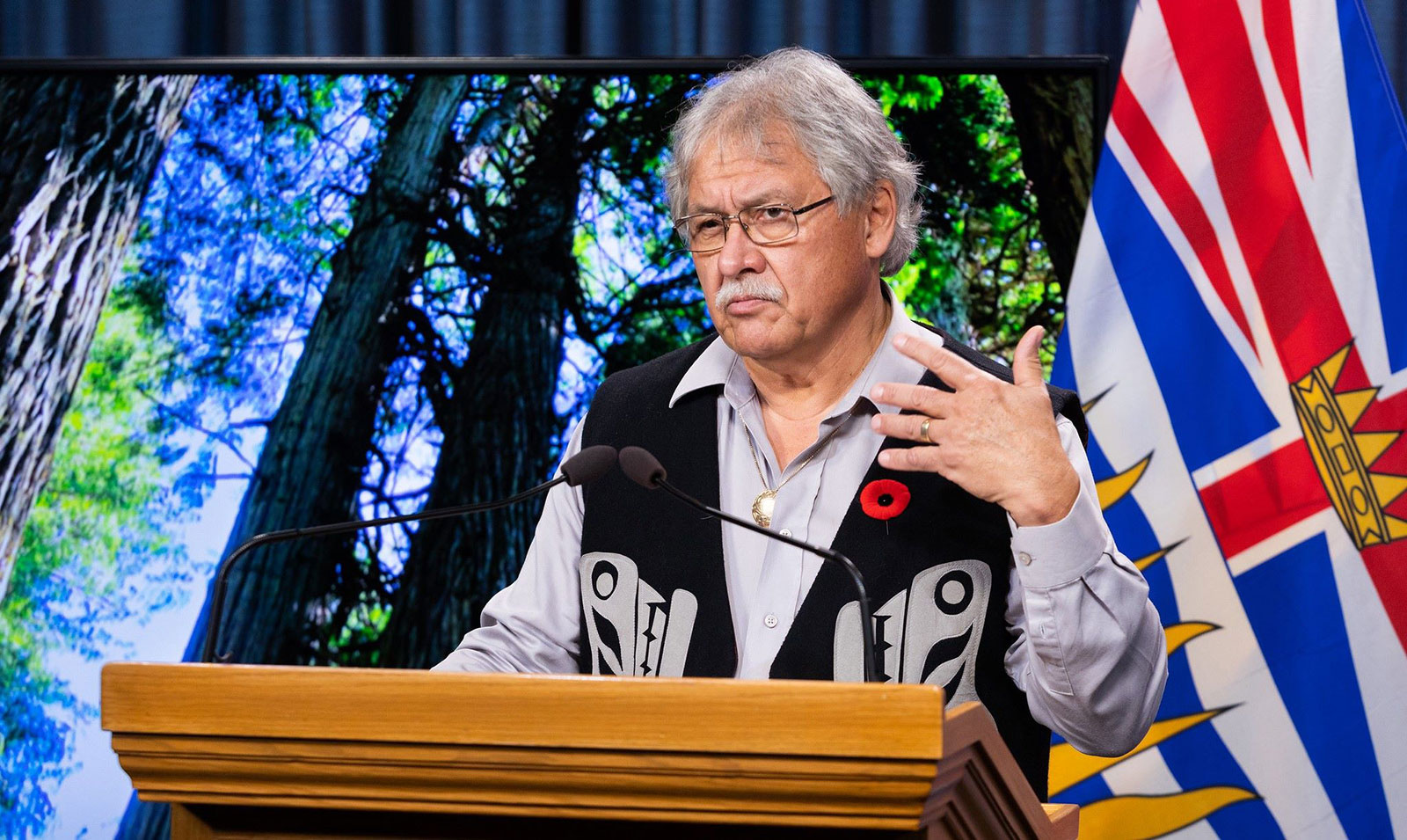 A silver-haired, medium-skinned man wearing a vest with Indigenous designs gestures from a podium.