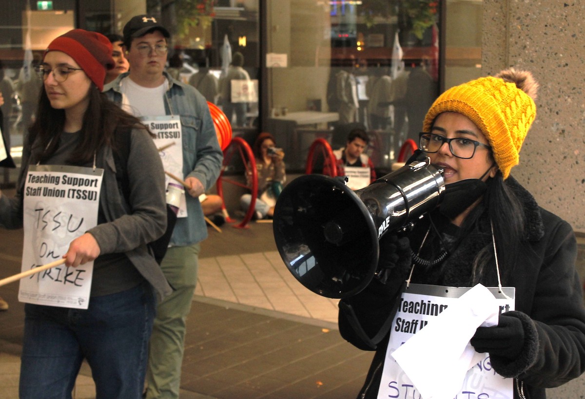 A woman wearing a yellow knit toque speaks into a megaphone, with a sign saying 'Teaching Support Staff Union' hung around her neck. To her right, another woman wearing a red toque wears a sign that says 'TSSU on Strike.'