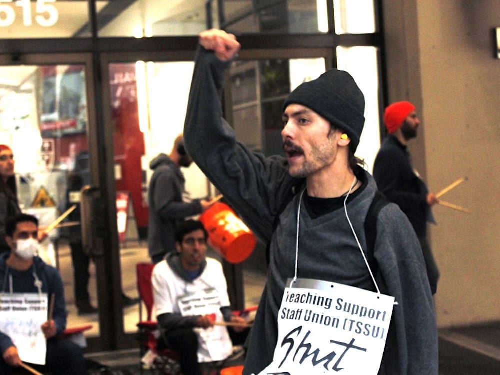 A man wearing a black toque holds a fist up in the air and wears a sign saying 'Teaching Support Staff Union' hung around his neck. In the background are other people wearing signs and holding drumsticks.