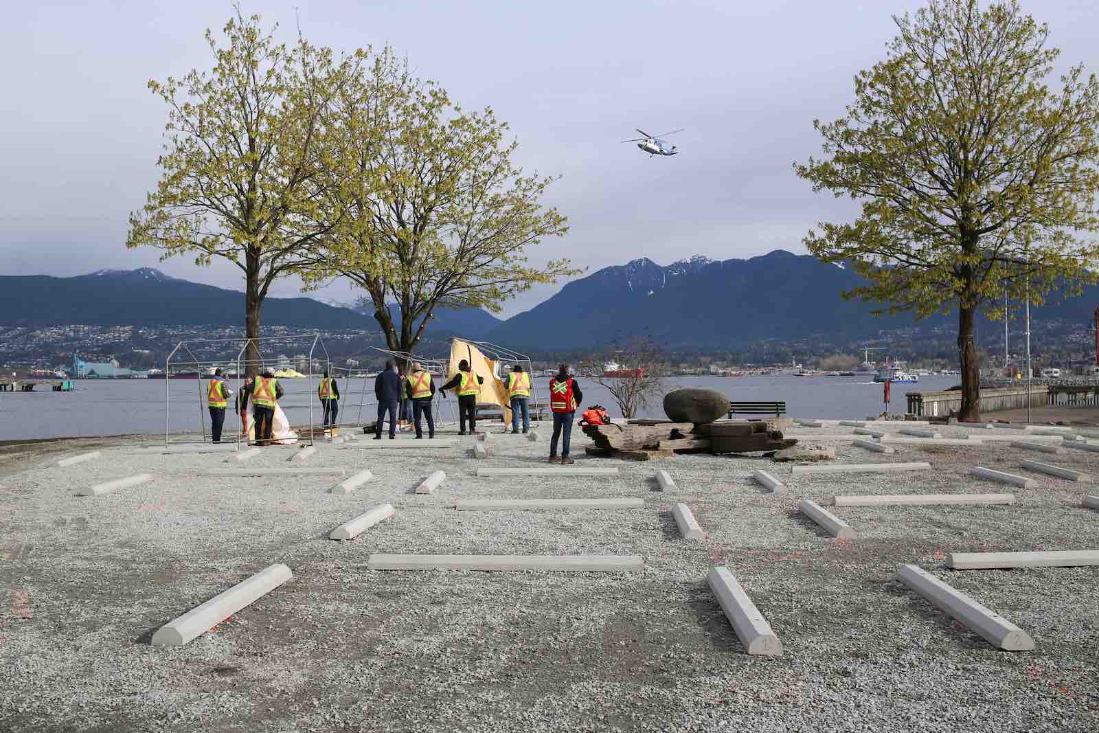About 10 workers in safety vests stand in a gravel-covered space with low concrete barriers marking tent sites. They are in front of Burrard Inlet.
