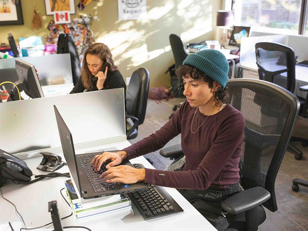 Two people work at laptop computers and phones in a brightly lit indoor office space. The person in the foreground has curly dark hair and a teal toque and is typing at a laptop. The person in the background has long dark hair and is holding a landline phone to their ear.
