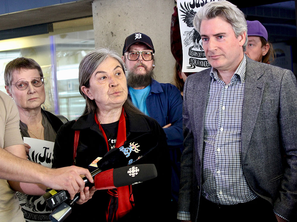 An Indigenous woman with long grey hair stands with microphones in front of her. A light-skinned man with grey hair wearing a grey jacket and dress shirt is beside her, and two more people are behind her.