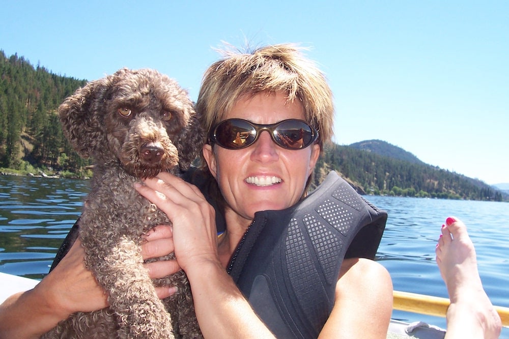 A smiling woman wearing sunglasses and a life jacket holds a light brown dog, with a lake behind her.