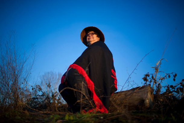 Judith Sayers is seated on a log against a blue sky. She is wearing a cedar hat and black and red blanket. She is looking towards the horizon.