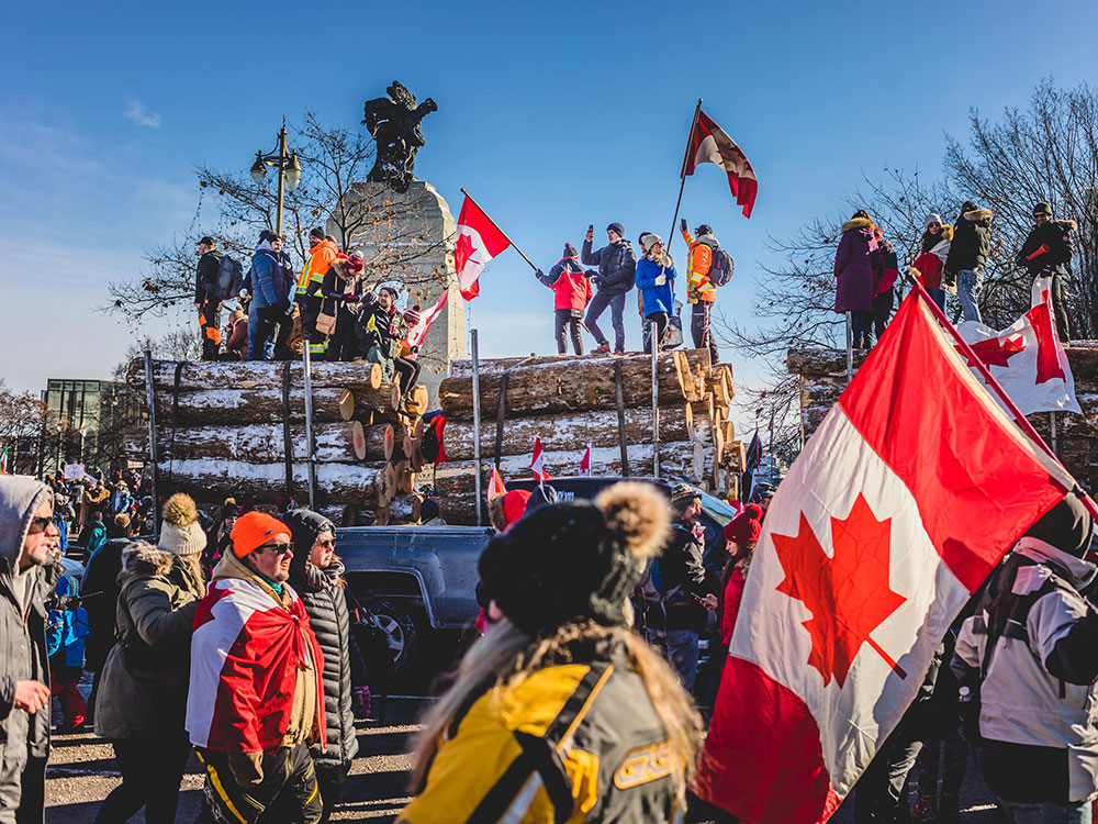 A scene from the Ottawa convoy protests. Protesters, some with Canadian flags, are walking in front of a truck carrying snow-covered logs.
