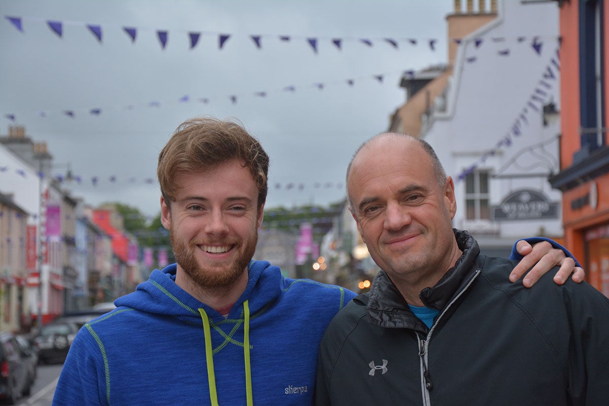 Two men stand in a festive urban street where purple bunting hangs across buildings. Kyle Fawkes, left, is wearing a blue hoodie. He has his arm around his father, Glen, right, in a black top. They are both looking at the camera and smiling.