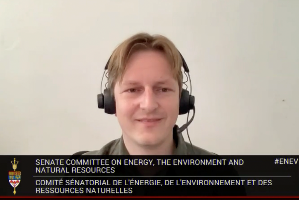 A man wearing over-the-head earphones and parted blond bangs looks at the camera in front of a beige background. A label reading “Senate Committee on Energy, the Environment and Natural Resources” spans the bottom border of the screen.