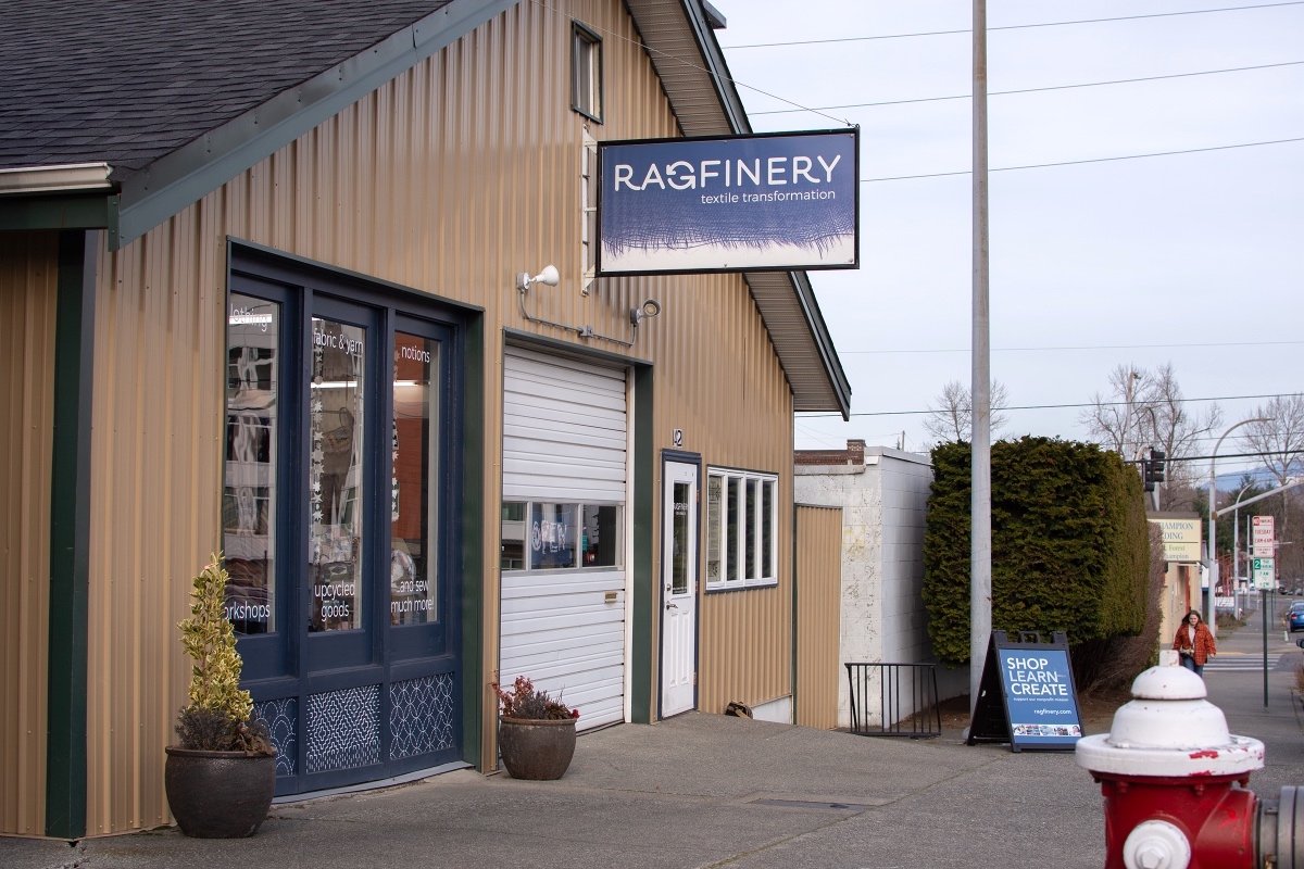 The exterior of the Ragfinery, a two-storey building on a Bellingham street with a peaked roof and beige vinyl siding with dark blue trim and a white roll-up garage door. A blue rectangular sign with the word 'Ragfinery' hangs from the second storey. It’s a dry, crisp winter day and the sky is light blue.