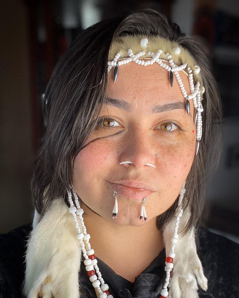 Close-up of Hanna Sholl, a medium-light-skinned person with short brown hair, hazel eyes and freckles. She is wearing beadwork around her face and has piercings through her nose and lower lip.