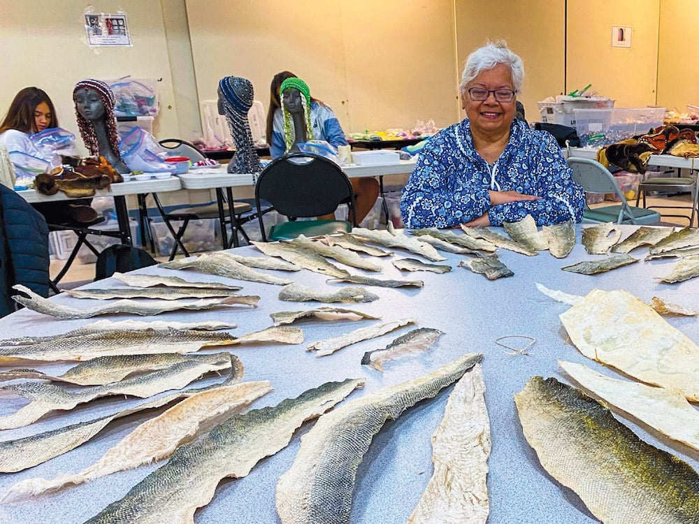 A woman with short white hair and glasses sits smiling at a table on which are spread out many salmon skins.