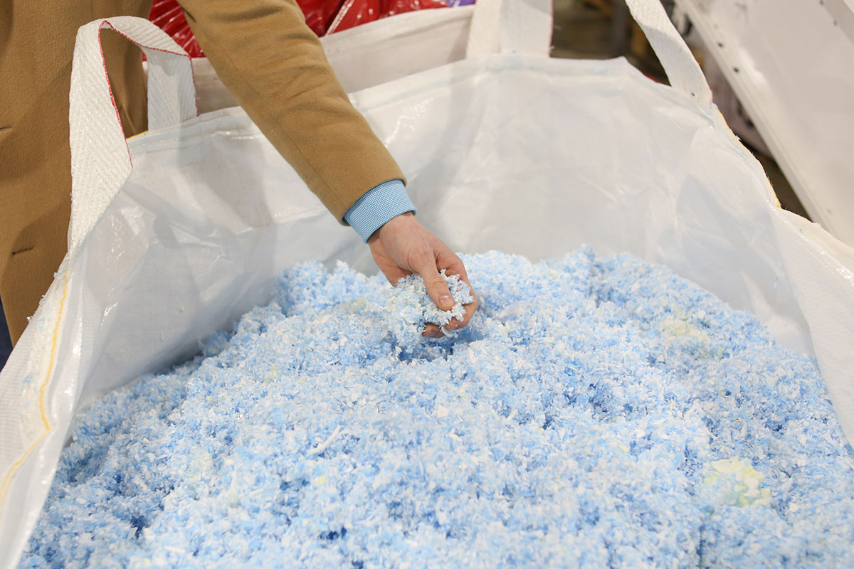 The hand of a light-skinned person reaches into a large white bag of light blue 'fluff' made from shredded polypropylene masks.