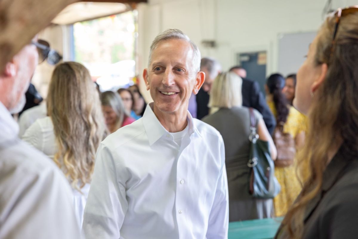A light-skinned man with short grey hair wearing a white dress shirt smiles while looking at someone to his right in a room full of people.