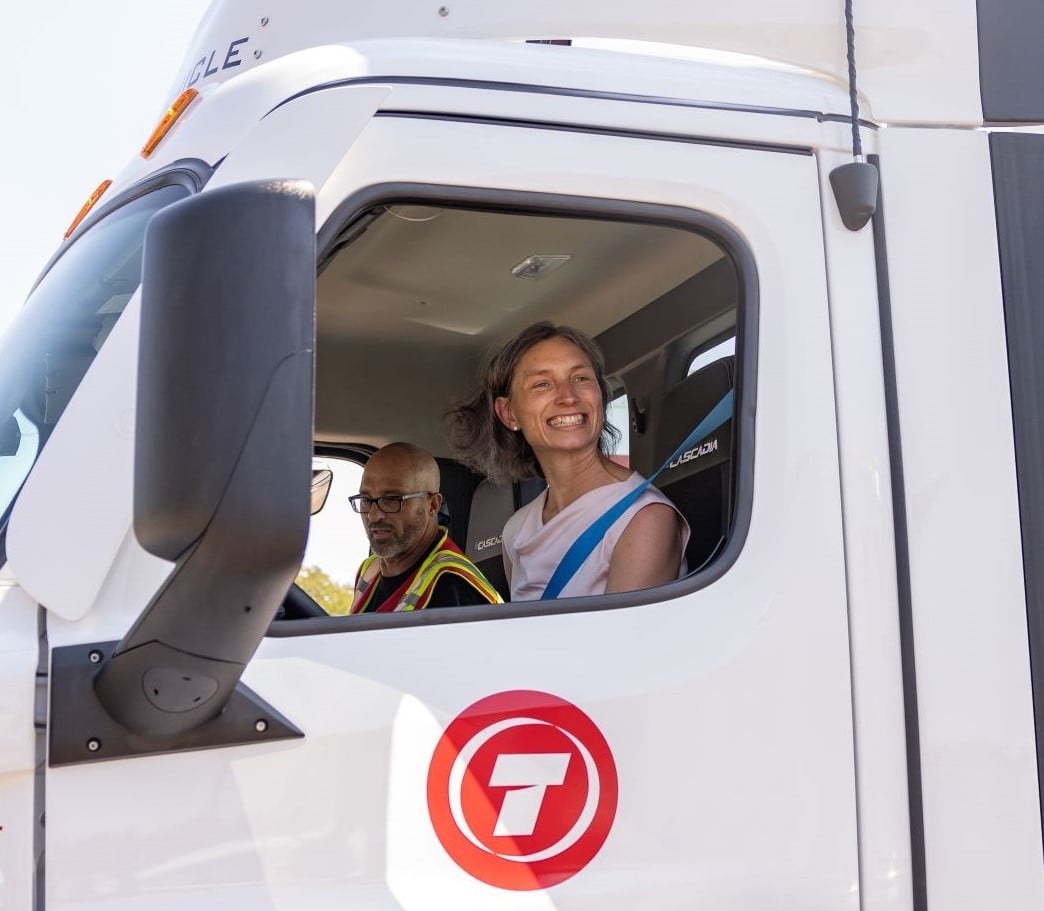 A smiling light-skinned woman sits behind the wheel in the cab of a white electric truck.