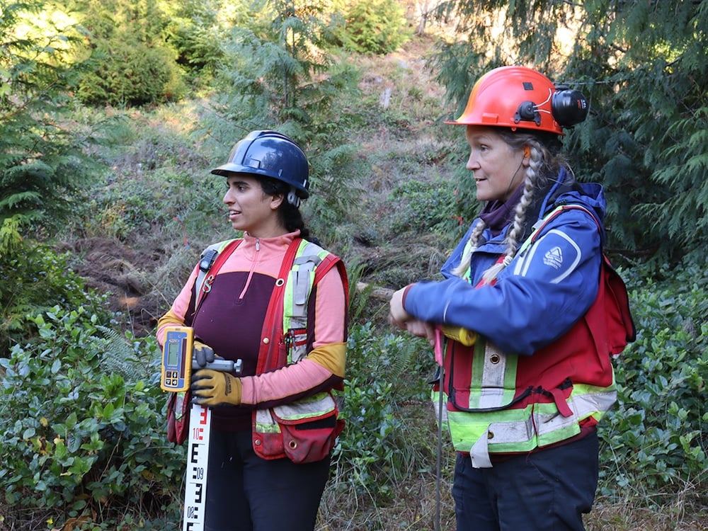 Two women wearing high-visibility vests and hard hats look off to the left.