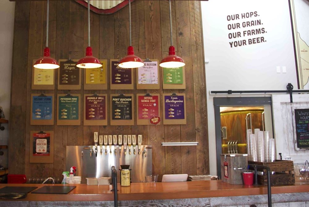 A row of four red pendant lights sits above the wooden bar at Barnside Brewing. Behind the bar is a steel tap and colourful papers on brown clipboards that feature the daily selection of beers.