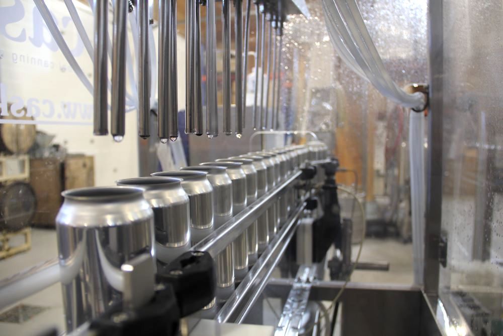 A row of tall silver beer cans sits under a row of spouts dispensing beer in a processing facility.