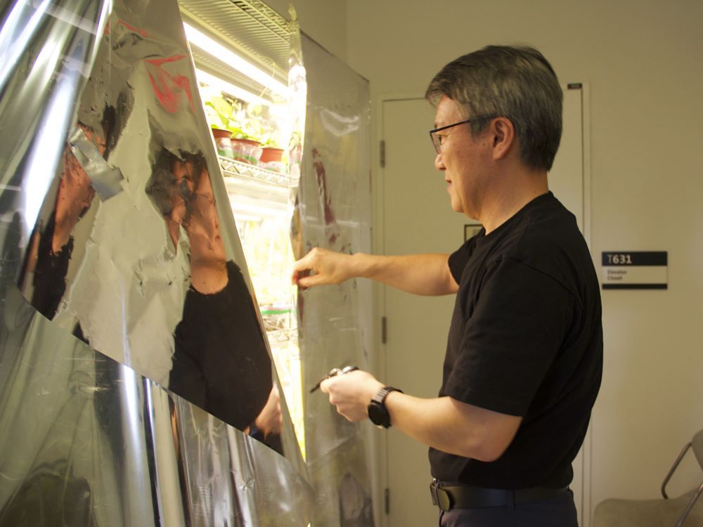 Ji Yang has short greying hair and glasses and is wearing a black T-shirt. He stands to the right of the frame and pulls back the silver curtain of a hydroponic shelf featuring rows of feral hop seedlings.