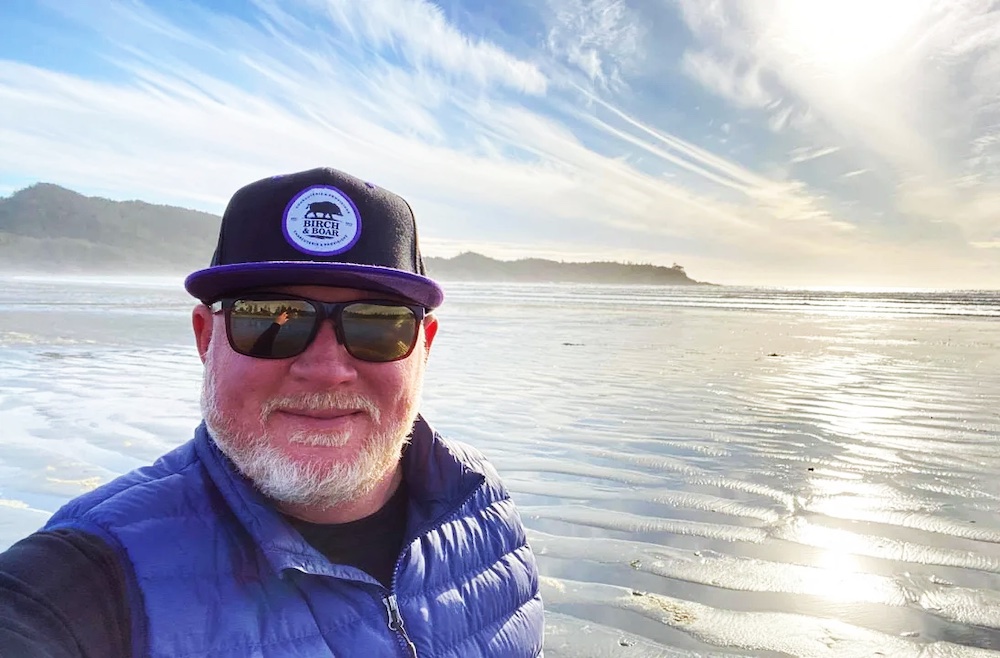 A bearded middle-aged white man in a ball cap smiles for a selfie taken on a beach on bright day, the sun glinting on the sand and water behind him.