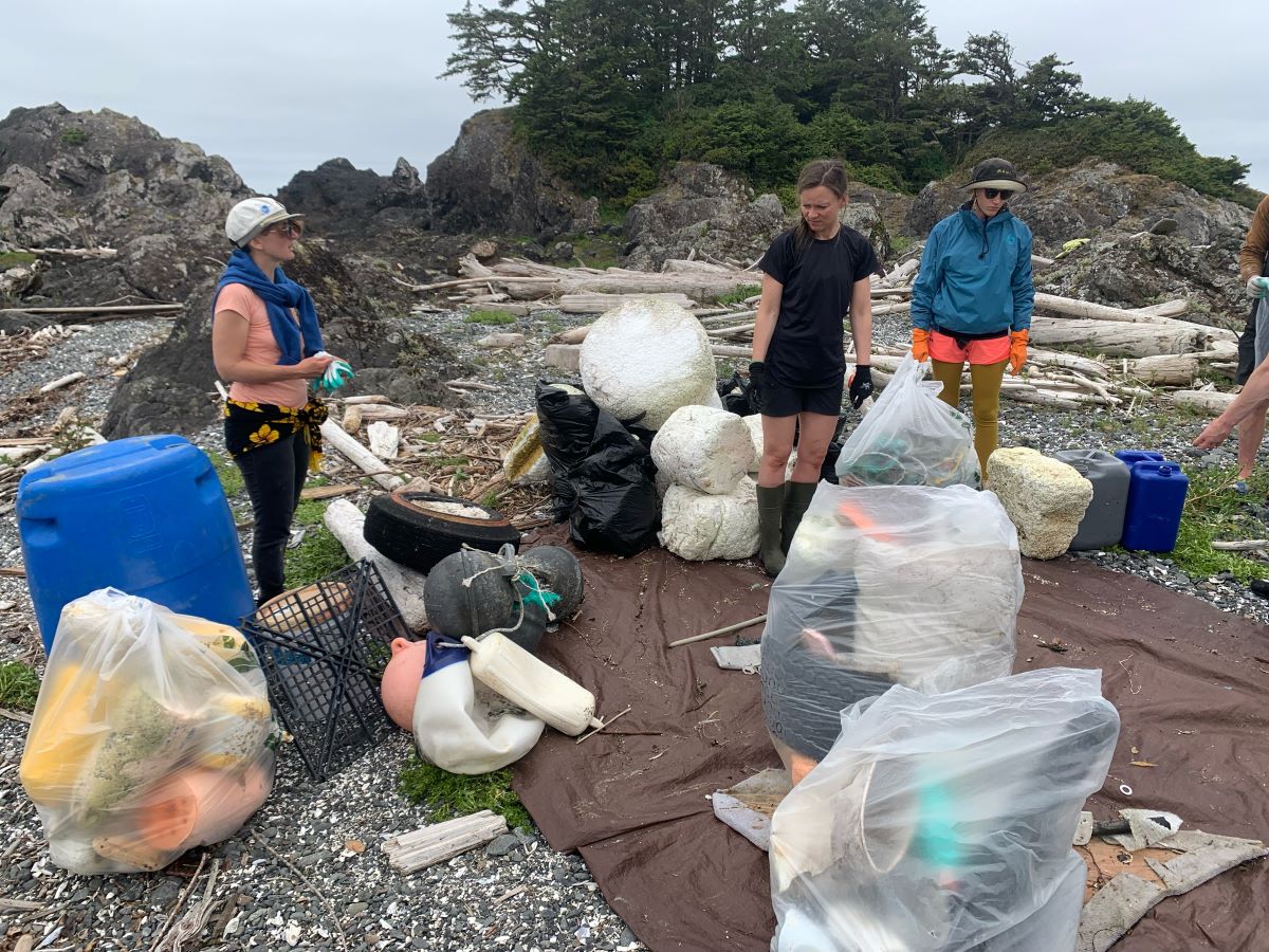 A group of people stand on a rocky beach with large plastic bags full of refuse.
