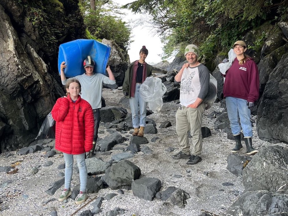 A group of cheerful people, some holding plastic refuse, stand on a pebbly beach with trees and large rocks around them.