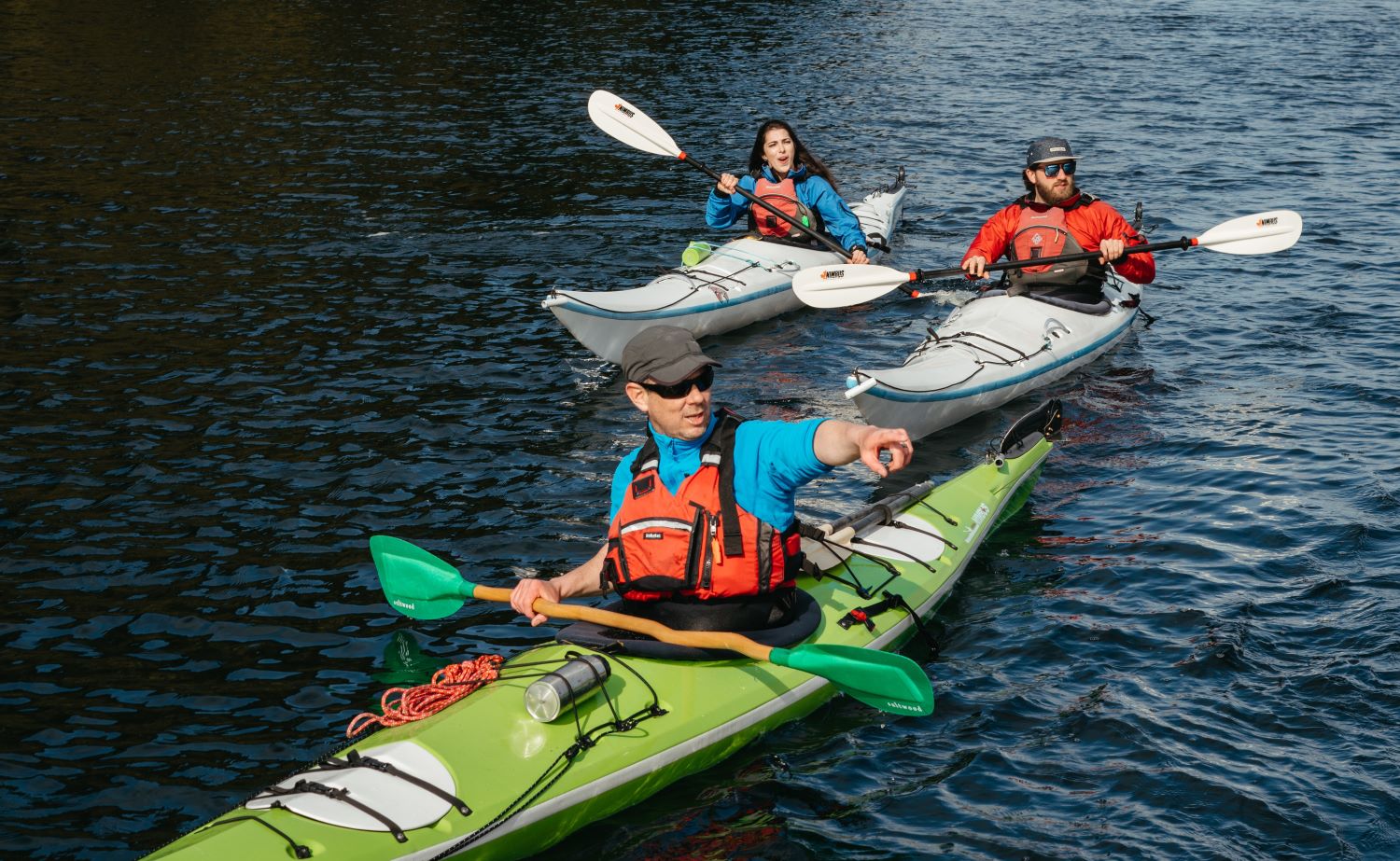 Two kayakers, a man and a woman, paddle in the ocean behind another kayaking man in a ball cap who points ahead.