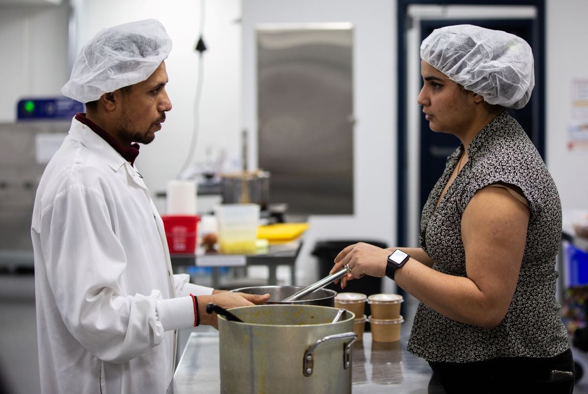 Two people, both with medium skin tone and wearing white hair nets, stand facing each other over pots in a kitchen. The man on the left wears a white chef's jacket. The woman on the right wears a short-sleeved patterned blouse.