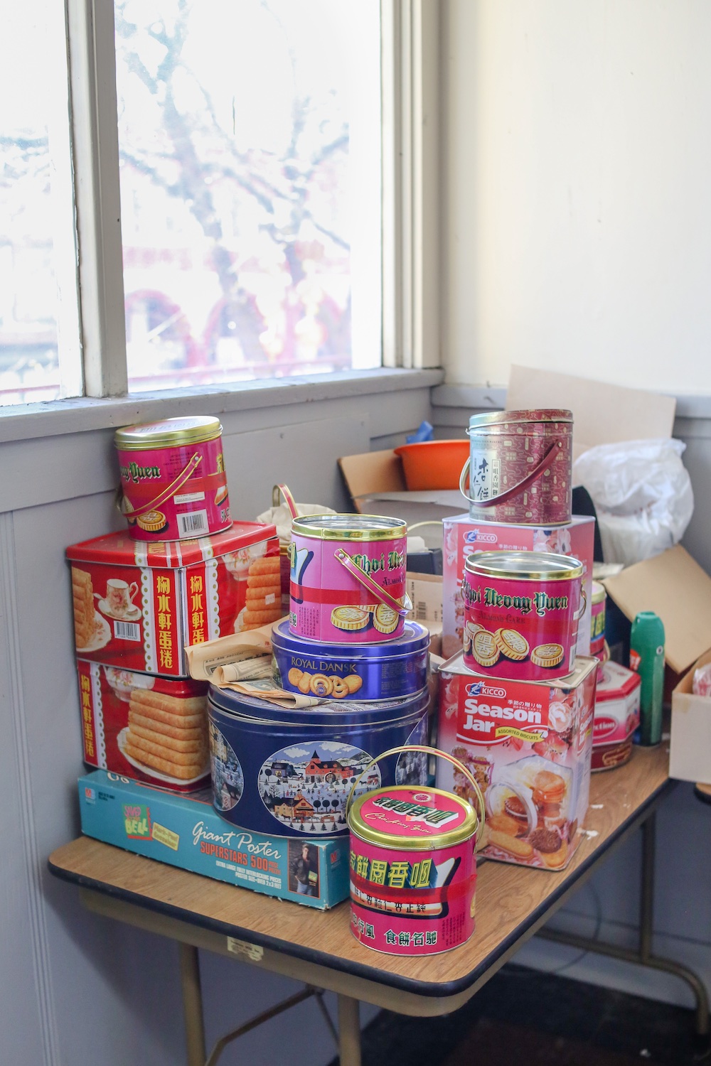 A stack of red, blue and pink cookie tins sits on an old wooden table loaded with discarded boxes and other packaging.