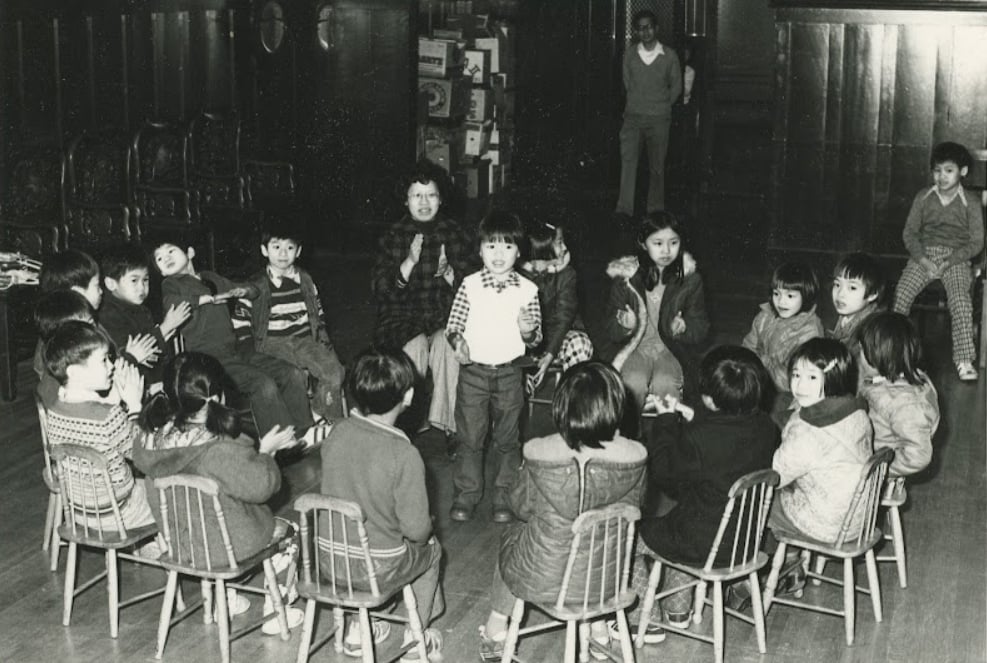 A black and white photo from the 1970s depicts children at the Mon Keang School for learning Cantonese. Young elementary school-aged children are seated in a classroom on small wooden chairs in a circle. Some are clapping while one child stands in the middle of the circle.