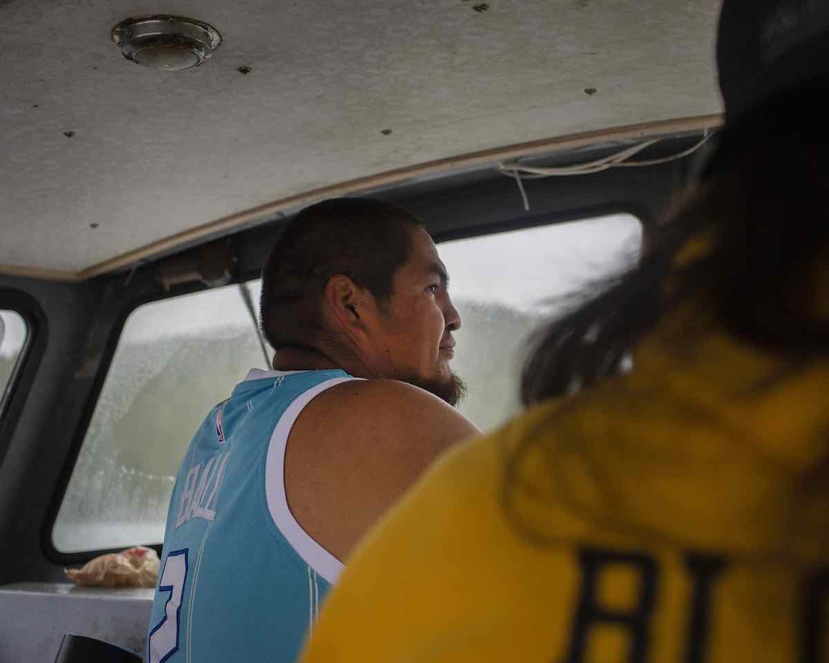 An Indigenous man with medium skin tone wears a sleeveless light blue basketball shirt. He faces the right of the frame and is indoors at the helm of a small boat. In the soft-focus foreground, a person wearing a yellow hoodie and long dark hair stands to the right of the frame. Their face is not visible.