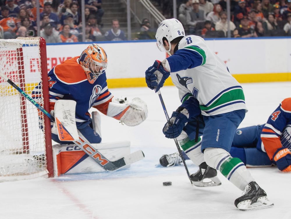 A Vancouver Canucks hockey player faces off with the goalie of the Edmonton Oilers in a rink with the audience watching closely in the background.