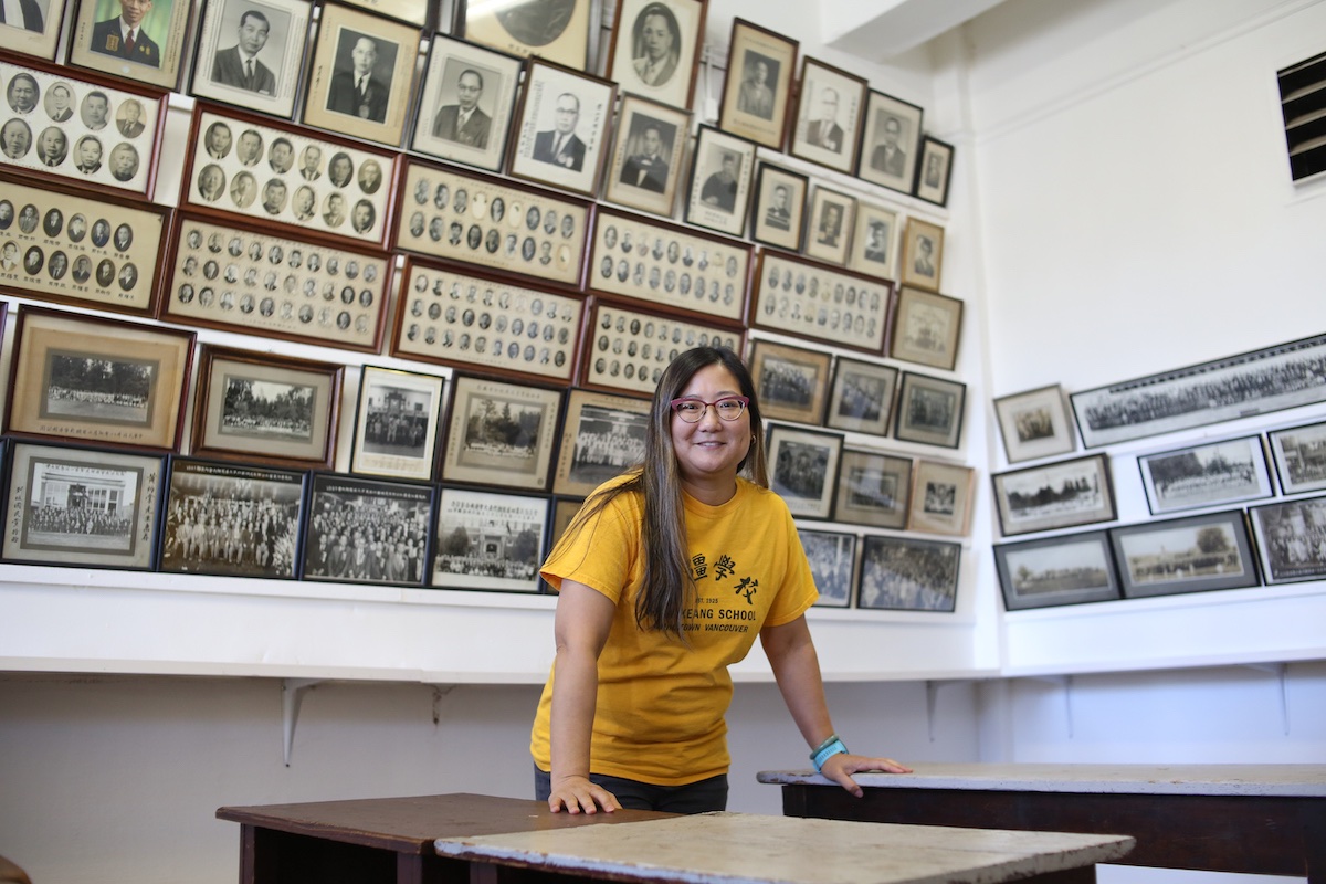 Aynsley Wong leans against a desk in front of a white wall full of framed black and white photos of Wongs’ Benevolent Association Society directors. She is a Chinese woman with long dark hair with blond highlights, glasses and a yellow T-shirt.