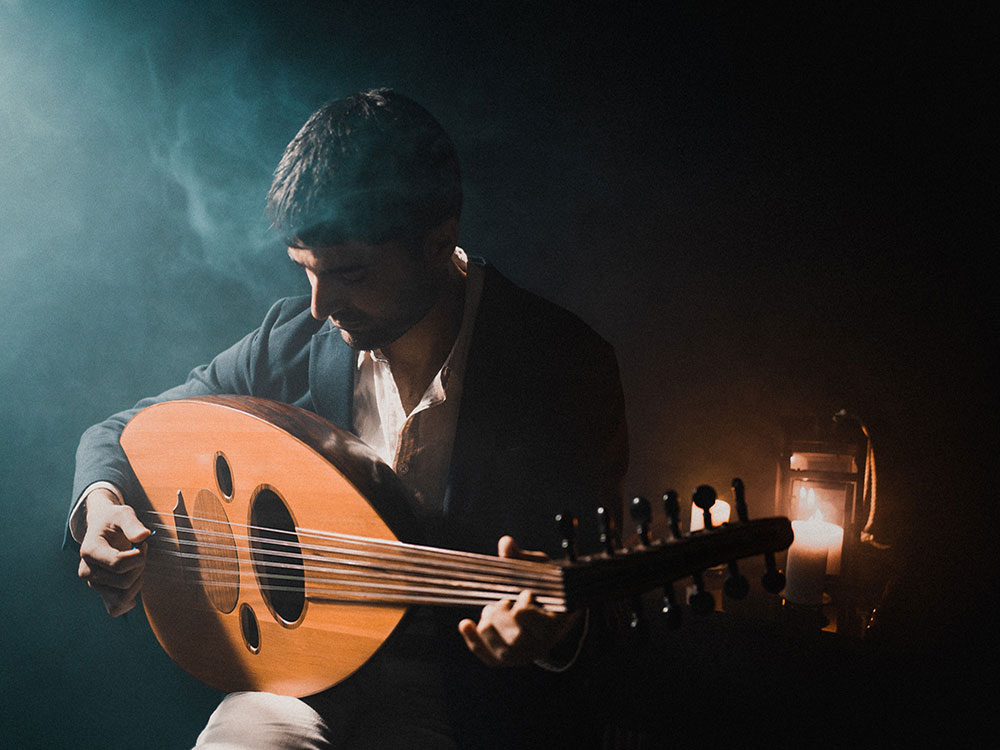 A man holding an aud looks down on the strings amidst a dark, smoky backdrop.
