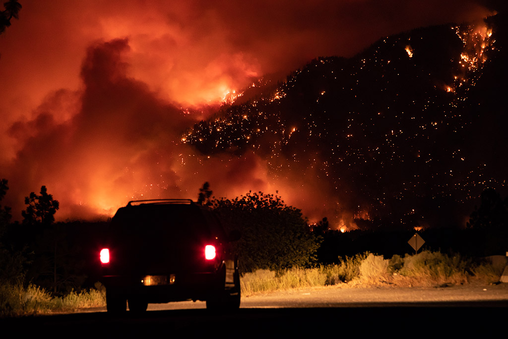 The sky is dark and fiery red. A driver has paused to take it all in. Their headlights illuminate an area of the road directly in front of them.