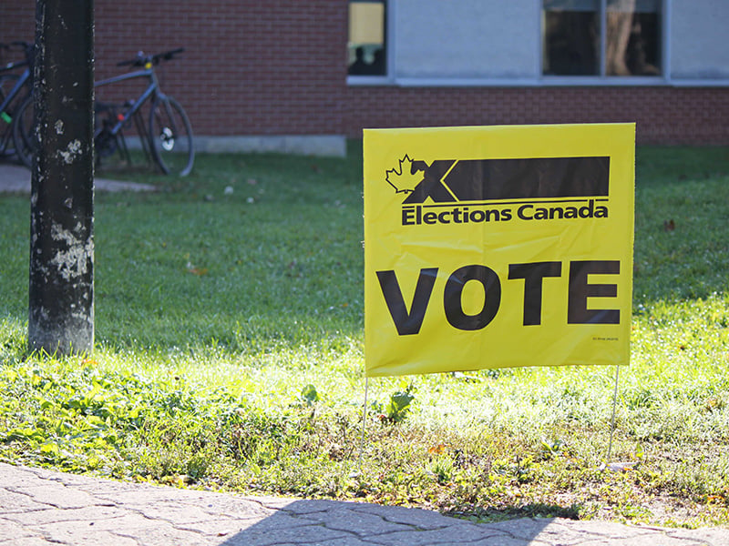 A yellow sign says, “ELECTIONS CANADA: VOTE.” It’s stuck into some green grass in front of a red brick building.  