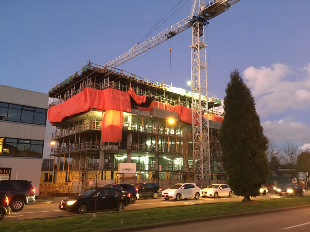 A crane arches over a six-storey building under construction along a Vancouver thorughfare. There is a dusky blue sky, cars on the road and lights on the worksite.