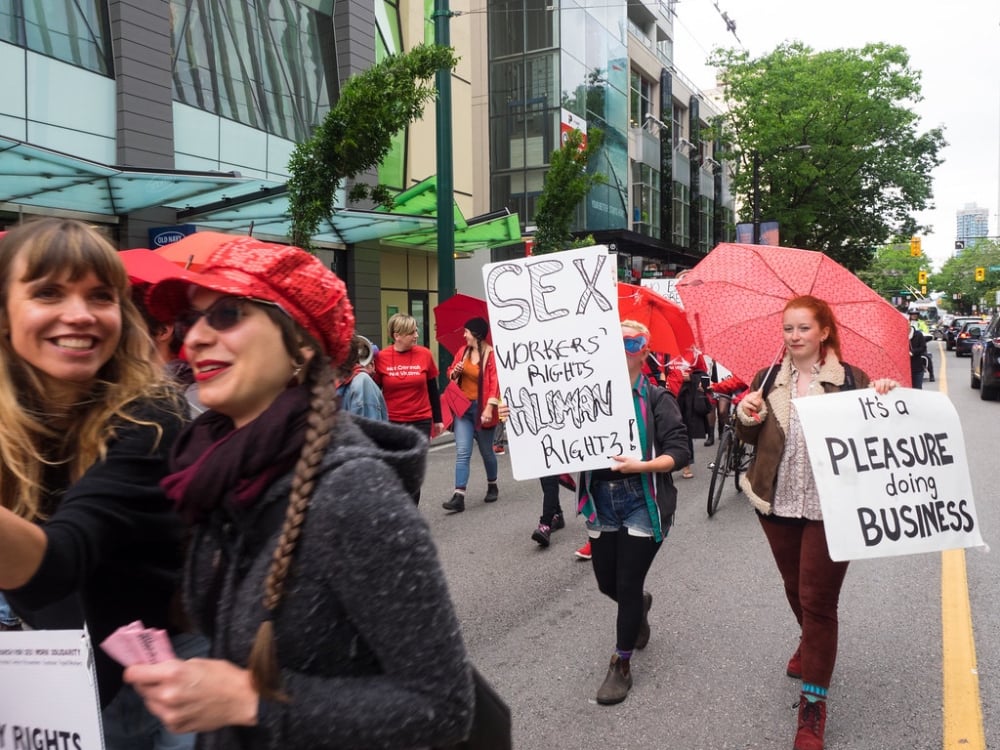 Women wearing red hats and carrying red umbrellas walk in a street protest. A sign reads “Sex Workers’ Rights, Human Rights.”