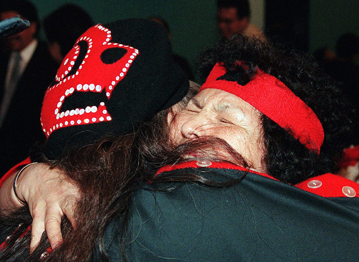 Two women wearing black and red Indigenous regalia hug tightly.