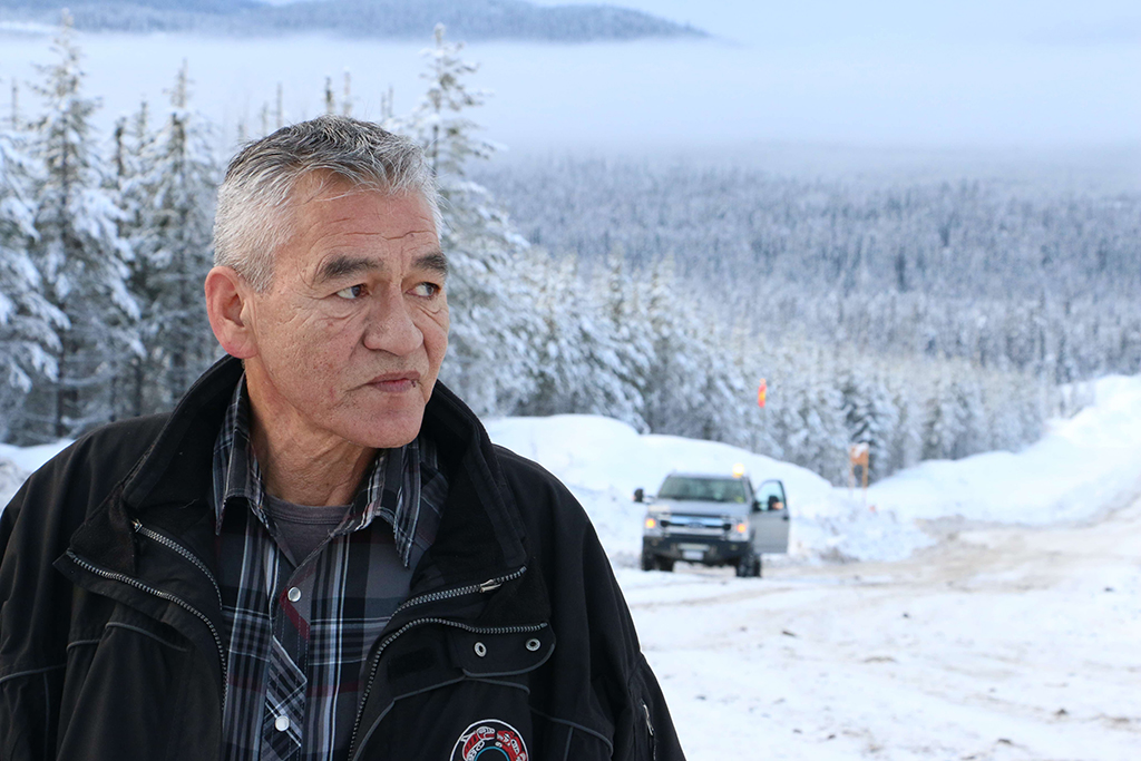 A middle-aged man with high cheekbones stand in a snowy landscape with a truck beyond.