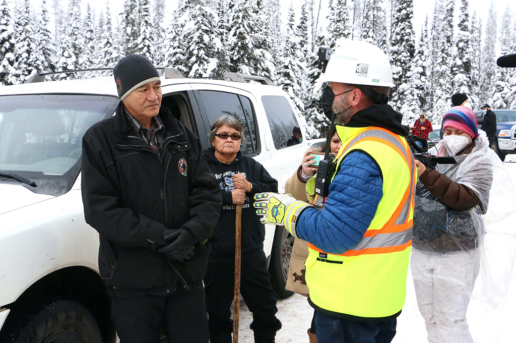 A man wearing a black coat and a black tuque, left, speaks with another man, wearing a mask and a yellow safety vest, right. There is snow on the ground and a few other people are standing near the men, witnessing. One person records with a video camera.