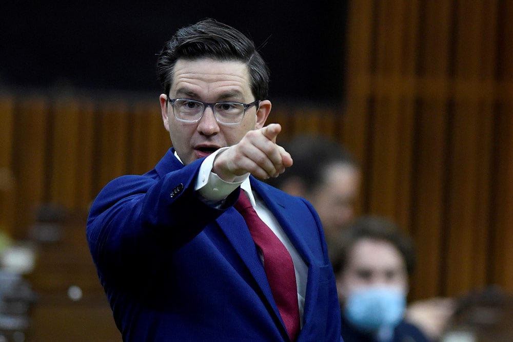 Pierre Poilievre, wearing a blue suit and red tie, points accusingly at someone out of frame.