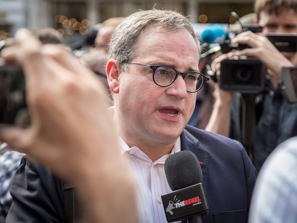Ezra Levant, holding a Rebel News-branded microphone, stands in the middle of a media scrum. Semi-blurred video news cameras surround him. 
