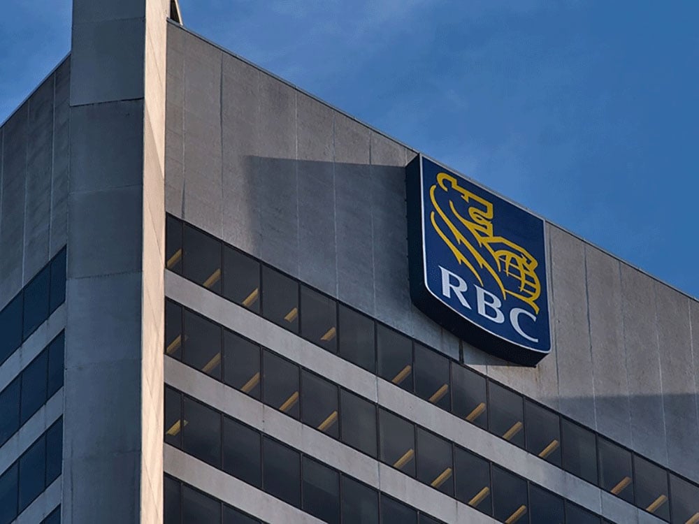 The top floors of a grey concrete office building against a blue sky, with a latge sign with the RBC logo of a golden lion clutching a goal and the letters RBC. 